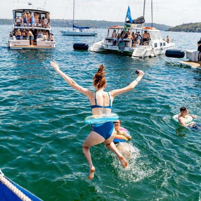 A person jumps into the water from a boat, wearing a blue swimsuit and an inflatable ring. Other people are swimming nearby, and several boats are anchored in the background under a partly cloudy sky.