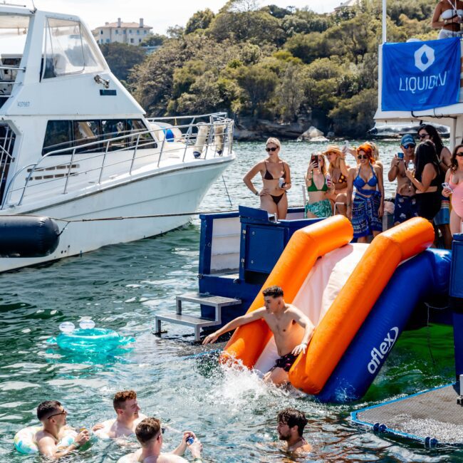 People are enjoying a boat party on the water. Some are on deck while others swim. A man is sliding into the water from a blue and orange slide. Another boat is nearby. The scene is lively and festive, with trees in the background.