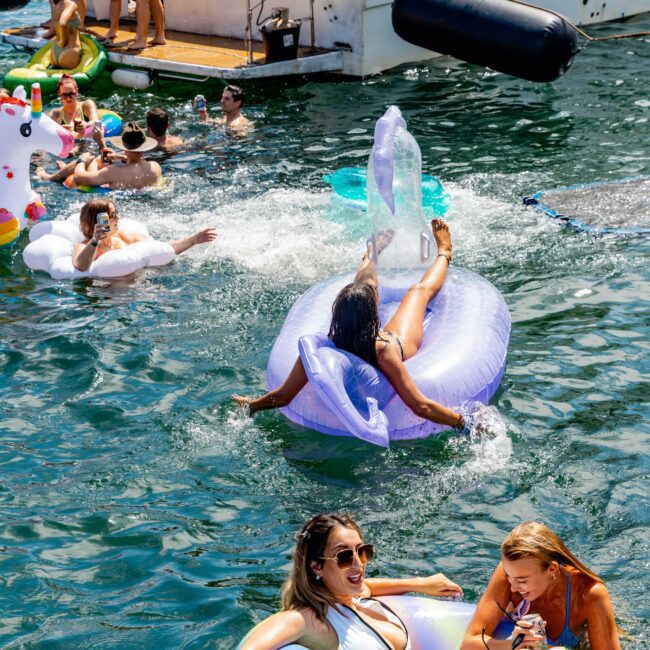 People enjoying a sunny day at a yacht party, swimming and lounging on colorful inflatable floaties in the water. A boat is docked nearby, with more people on board. The atmosphere is lively and vibrant.
