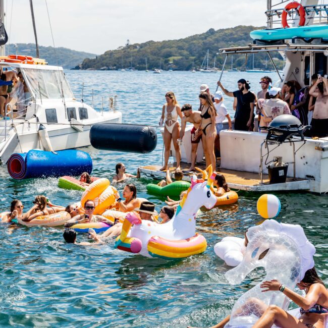 People enjoying a sunny day on floats and in the water next to boats. Some are on colorful inflatable toys like a unicorn. A group is on a boat deck, and the background shows a tree-lined shore and other vessels.