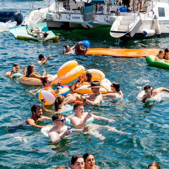 A group of people enjoying a sunny day in the water, surrounded by inflatable pool toys, near a docked boat. The scene is lively, with several colorful floaties and swimmers laughing and relaxing.