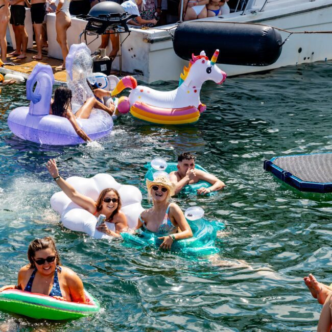 People enjoying a sunny day in the water near a docked boat. Some are floating on inflatable toys, including a unicorn and a swan. There's a happy, lively atmosphere as they relax and socialize.