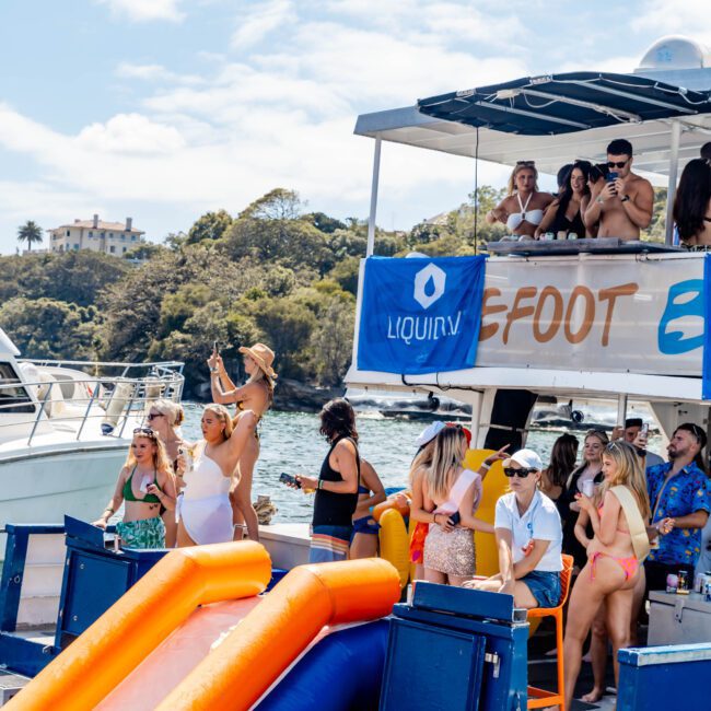 People enjoying a party on a boat with an inflatable slide into the water. Some are sitting, others are taking photos, and a few are dancing. The scene is festive with a clear sky and trees visible in the background.