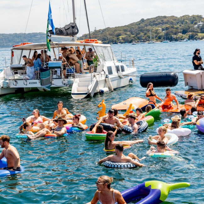A large group of people enjoying a sunny day on a lake, most floating on colorful inflatable tubes and rafts. A nearby boat is filled with more people, and there’s a dock where additional people gather. Trees line the background.