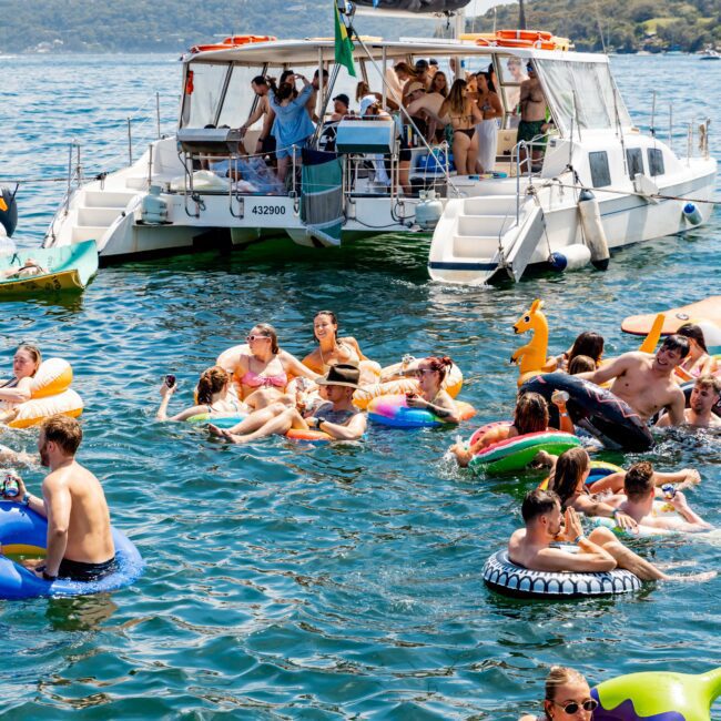 A group of people enjoying a sunny day on a lake. Many are on colorful inflatable tubes in the water, while others are on a large boat. Trees are visible in the background. The water is clear under a blue sky.