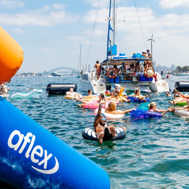 Groups of people on colorful floaties are enjoying a sunny day in the water near a large boat. A bridge is visible in the background. A large inflatable with the word "aflex" is in the foreground.