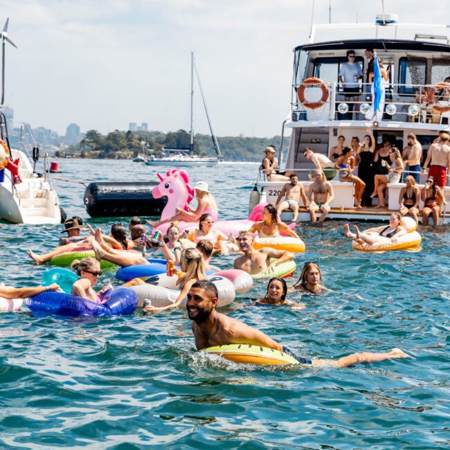 People enjoying a sunny day on the water, swimming and floating on colorful inflatables near a boat. A man swims nearby, and others socialize on the boat deck. The backdrop features a distant skyline under a partly cloudy sky.