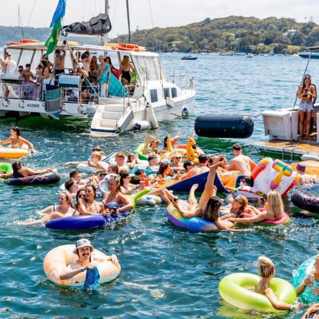 A lively group of people floating on inflatable rafts and rings in a sunny harbor, with a boat and land in the background. They are enjoying a festive atmosphere, some holding drinks. The water is clear and the sky is bright.
