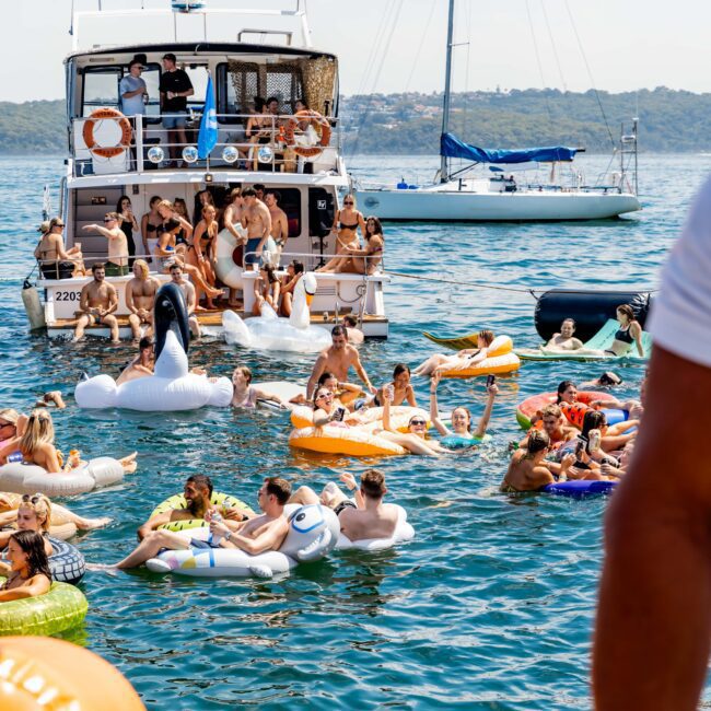 A sunny outdoor scene featuring a large group of people on inflatables in the water near a yacht. Some are lounging on colorful floatation devices, others are swimming. A sailboat is visible in the background. The atmosphere is lively and fun.