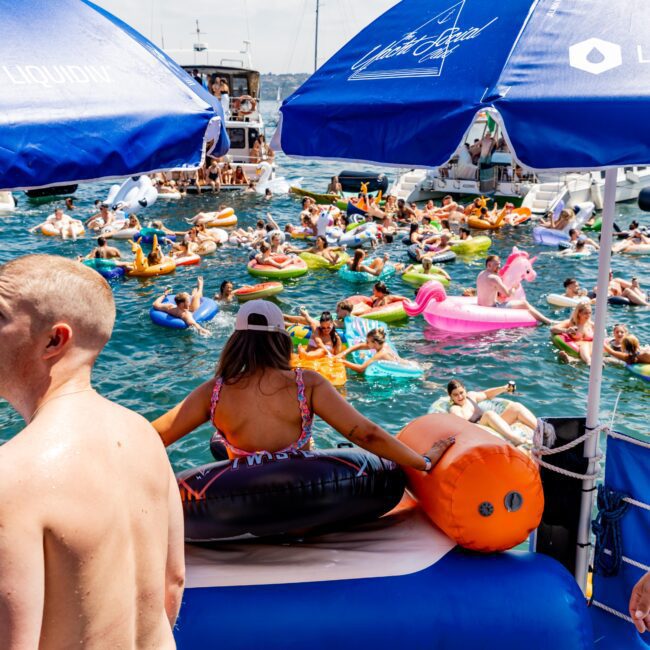 A large group of people enjoying a sunny day on the water, floating on colorful inflatable tubes and rafts, including a unicorn. Blue umbrellas provide shade on a boat deck in the foreground.