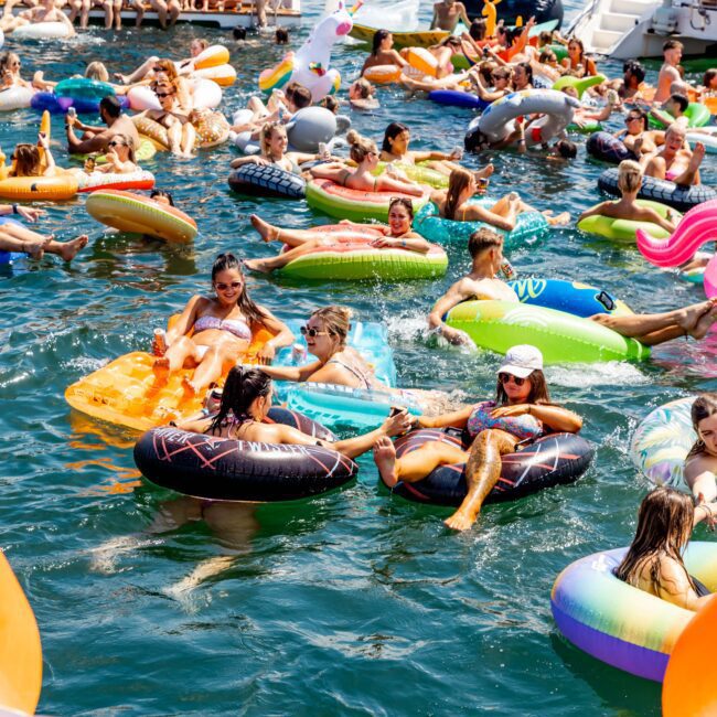 A lively scene of people enjoying a sunny day on the water with colorful inflatable floats. The water is crowded with various inflatables, and boats are visible in the background. People are relaxing and socializing, creating a festive atmosphere.