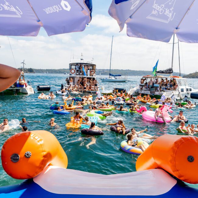A large group of people enjoying a sunny day on the water with colorful inflatables and boats. Two umbrellas on a boat frame the scene, and various floaties including swans and flamingos are visible. The text "Yacht Social Club" is in the corner.