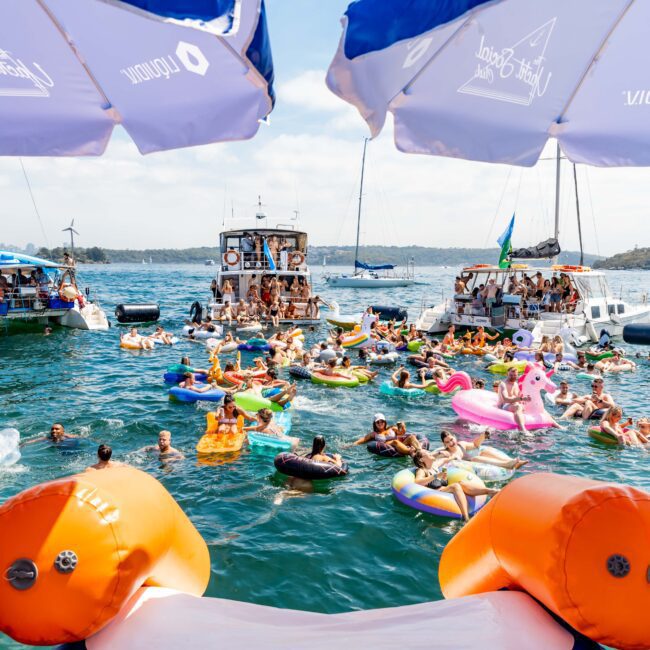 People enjoying a lively yacht party in a bay, with many swimming and using colorful inflatables. Several boats are moored nearby. The scene is lively, with umbrellas and buoys in the foreground and lush hills in the background.