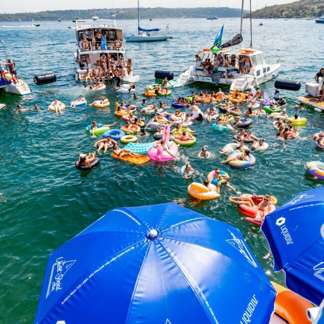 People enjoying a sunny day on the water, surrounded by yachts and boats. Many are floating on colorful inflatables, and a large blue umbrella in the foreground reads "The Yacht Social Club". The backdrop features distant hills and a clear sky.
