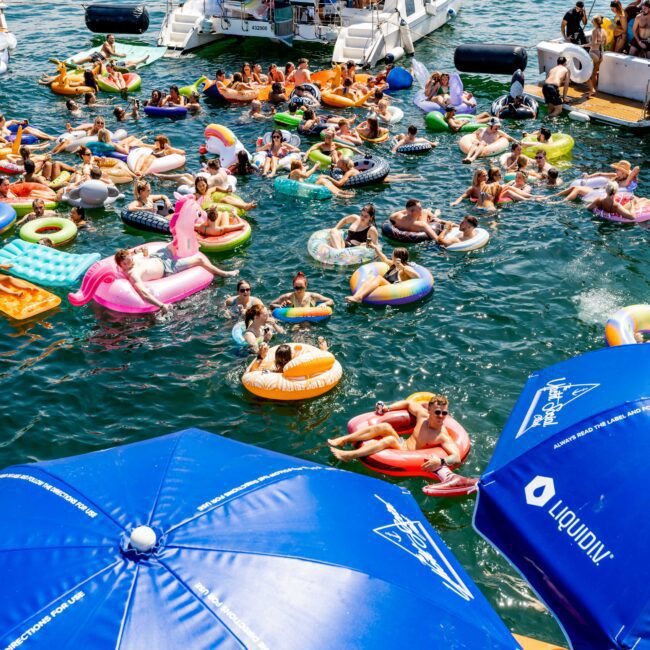 A large group of people enjoy a sunny day on a lake, floating on colorful inflatables near a boat. Blue umbrellas with logos are visible in the foreground, and several boats are anchored nearby.