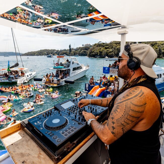 A DJ with headphones and sunglasses is performing on a boat deck. In the water below, a crowd of people on inflatable floats are gathered, enjoying the music. Boats and a scenic shoreline are visible in the background.