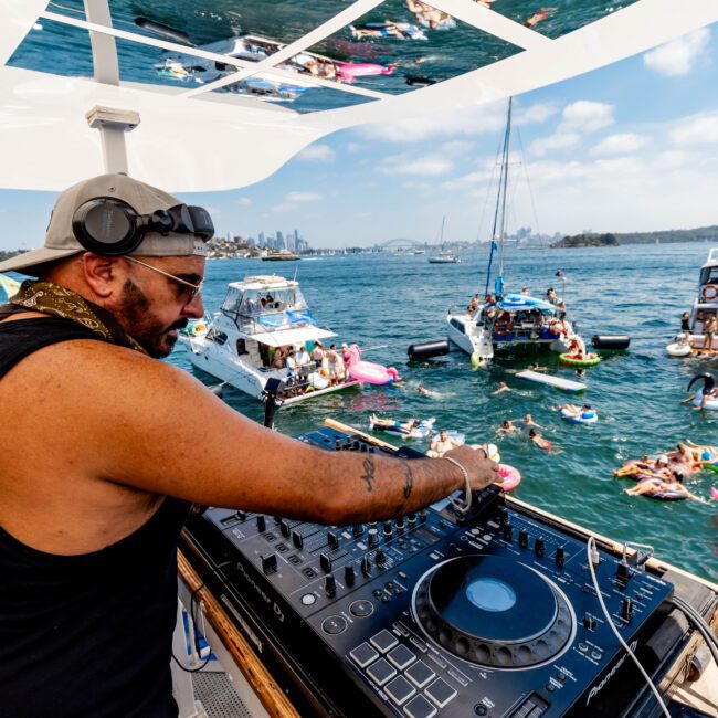 DJ playing music on a boat with a mixer, wearing headphones and a sleeveless shirt. Reflections of people on inflatables in the water are seen above. Other boats and swimmers are visible in the background.