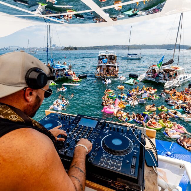 DJ playing music on a boat with turntables. People on colorful inflatables are enjoying the water, surrounded by other boats. The background features a city skyline and bridge under a clear blue sky.