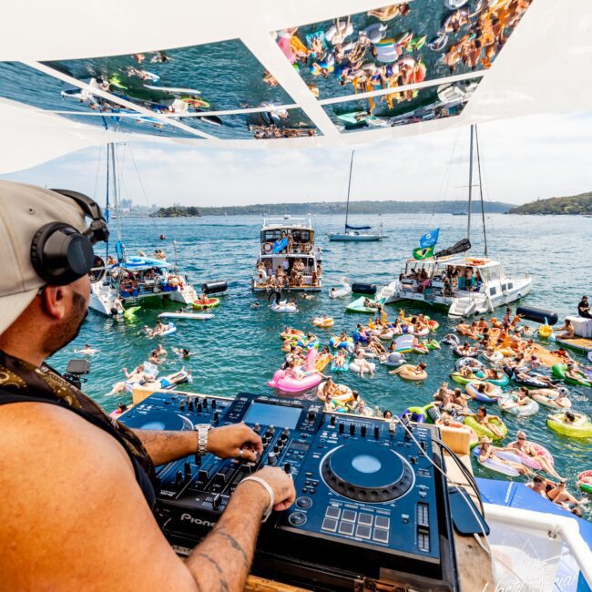 DJ playing music on a boat with a crowd of people on inflatable rafts and boats enjoying a sunny day on the water. The scene is lively with people swimming and socializing, surrounded by yachts and sailboats.