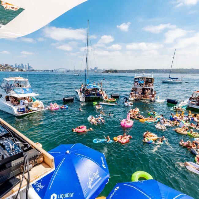 Boats are anchored in a bay with people relaxing on colorful inflatable floats in the water. A DJ setup with turntables is on one boat, suggesting a lively party atmosphere. Clear skies and a distant city skyline enhance the festive scene.