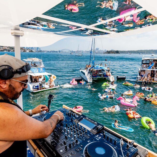 DJ performing on a boat deck with a mixer, while people in swimwear relax on inflatable floats in the water. Several boats and a city skyline are in the background.