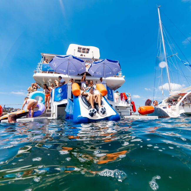 People enjoying a sunny day on a yacht with a water slide, surrounded by inflatables and clear blue sky. Swimmers can be seen in the water near the slide.
