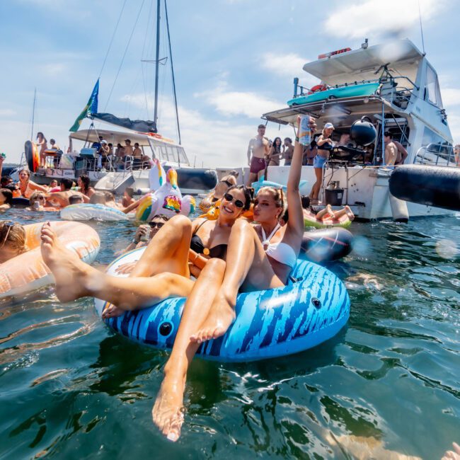 A group of people enjoying a sunny day on the water. Two individuals sit on a blue inflatable, surrounded by others on floaties and boats. The background features yachts and a bright blue sky. The atmosphere is lively and festive.