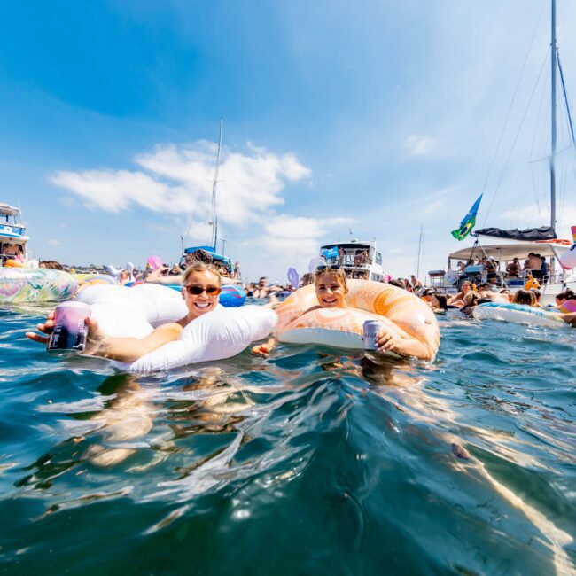 Two people float on inflatables in the water, surrounded by boats and other floaters. The scene is sunny with a clear blue sky. The atmosphere is festive and relaxed.