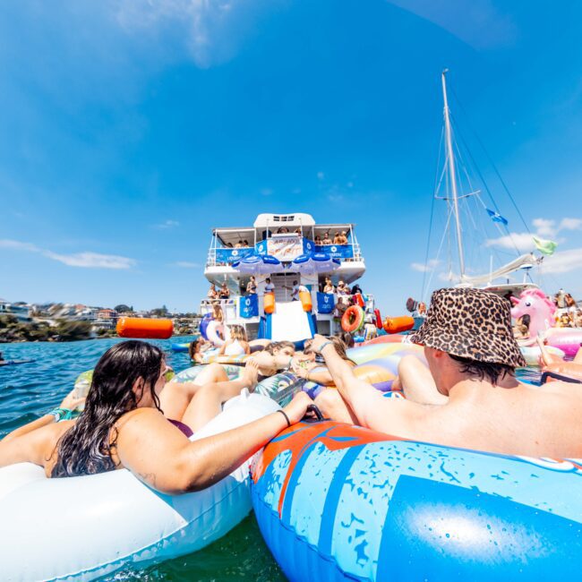 People are lounging on colorful inflatables in the water, with a large boat in the background. The scene is bright and sunny, and some trees and a sailboat are visible in the distance.