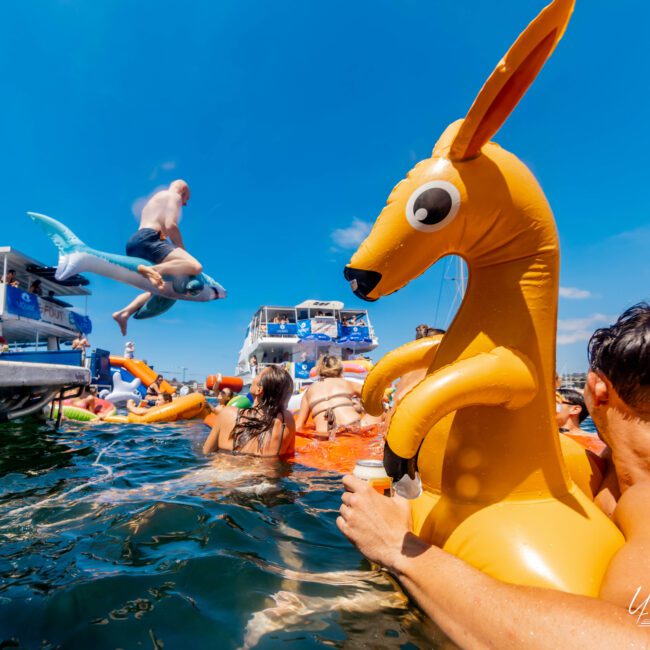 A lively boat party with people swimming and lounging on various inflatable pool toys, including a kangaroo. A person jumps off a boat with an inflatable dolphin. The sky is clear and blue, and several boats are anchored nearby.