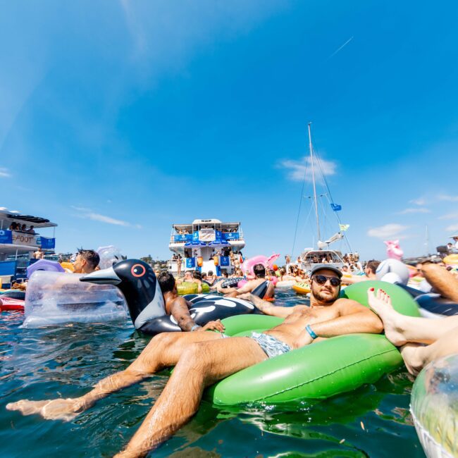A man relaxes on a green inflatable float in clear blue water. Surrounding him are other people on inflatables, a large boat, and a sunny sky overhead. The atmosphere is lively and festive.
