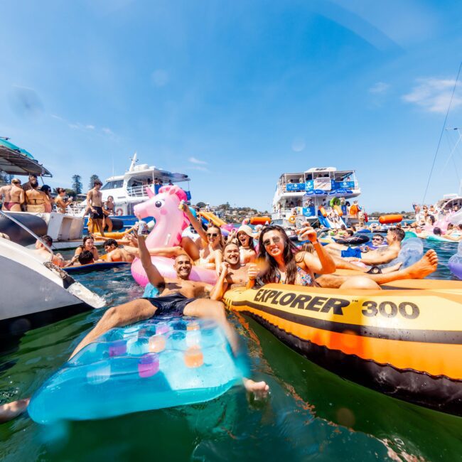 A group of people enjoying a sunny day on a lake, floating on inflatable rafts, including an orange raft and a pink unicorn. Boats and other inflatables are in the background. It's a lively, festive scene with clear blue skies.