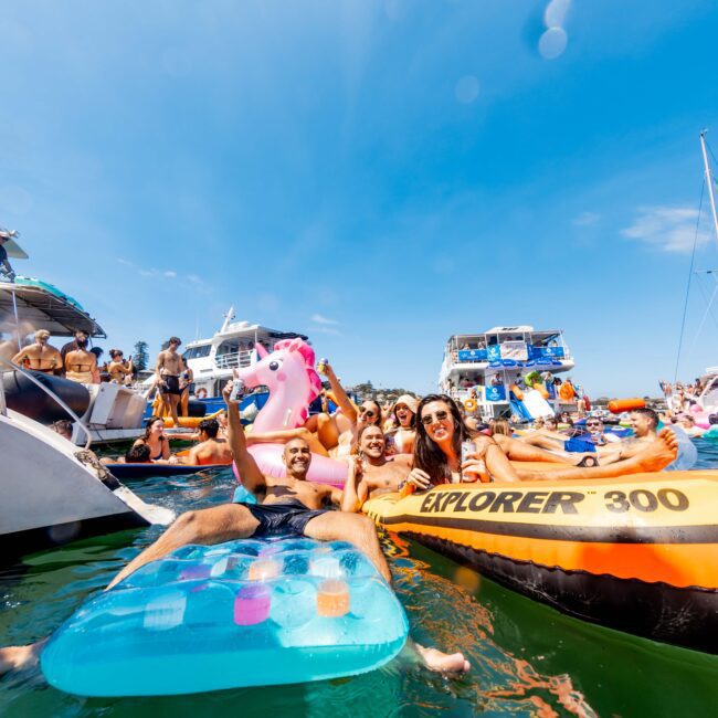 A group of people on inflatable floats enjoy a sunny day on the water, surrounded by boats and other party-goers. One person holds a pink inflatable unicorn. The scene is festive and lively, with a clear blue sky overhead.