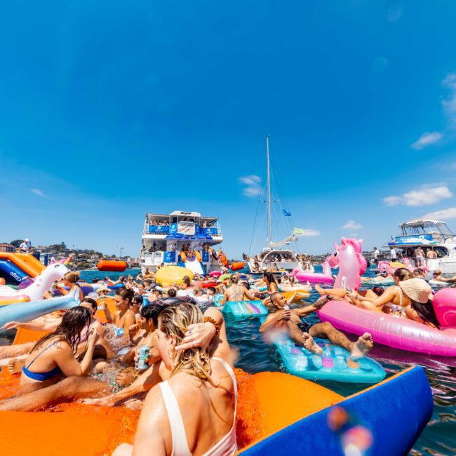 A bustling scene at a floating party with people relaxing on colorful inflatable rafts, including a unicorn. Boats are anchored nearby under a clear blue sky. Sunlight reflects on the water, creating a lively and festive atmosphere.
