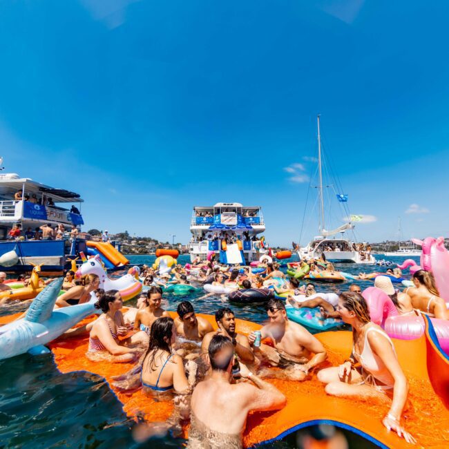 Crowd of people enjoying a sunny day on a lake, surrounded by colorful inflatables and boats. Some are in the water, while others relax on rafts. The sky is clear and blue, creating a festive and vibrant atmosphere.