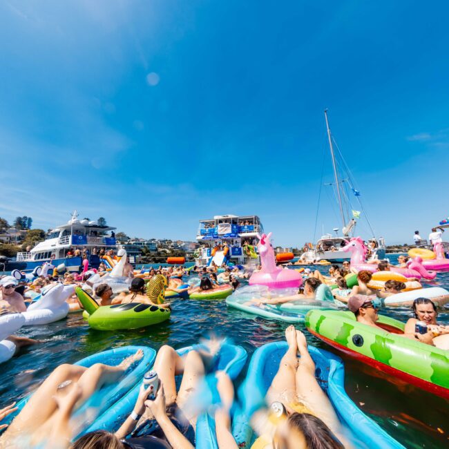 Crowd of people lounging on colorful inflatable floats in a sunny, blue-water setting, surrounded by boats. It’s a lively atmosphere with clear skies and numerous participants enjoying the day.