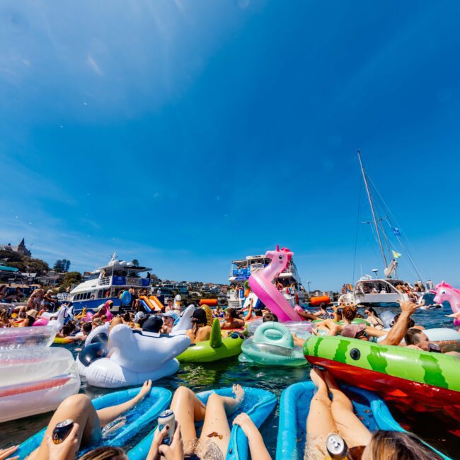 A lively scene on the water with people relaxing on colorful inflatable floats. Boats and a blue sky are in the background, while attendees hold drinks and enjoy a sunny day, creating a festive atmosphere.