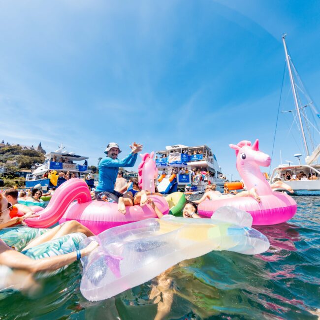 A lively scene on the water with numerous people enjoying a sunny day on inflatable rafts. Among them are pink unicorn and flamingo floats. Boats are docked in the background under a clear blue sky.