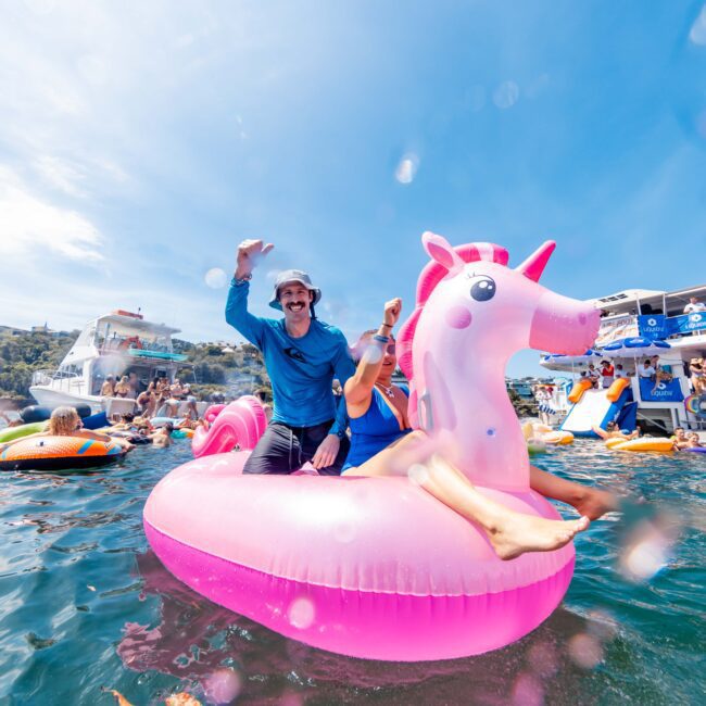Two people riding a large pink unicorn float on the water, surrounded by other people on floats. The background features boats and a clear blue sky. One person wears a hat and waves, while others enjoy the sunny day.
