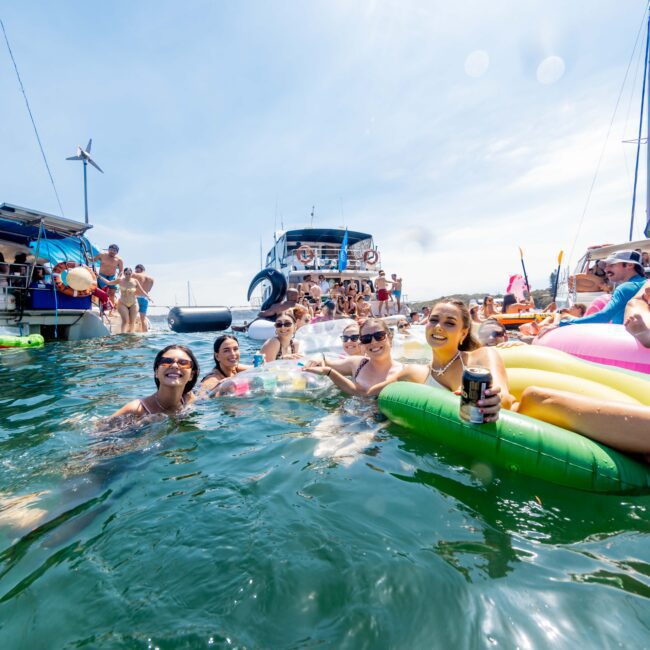 A group of people enjoying a sunny day in the sea, surrounded by boats. Some are swimming while others relax on inflatable floats, including a pink unicorn. The scene is lively with blue skies overhead.