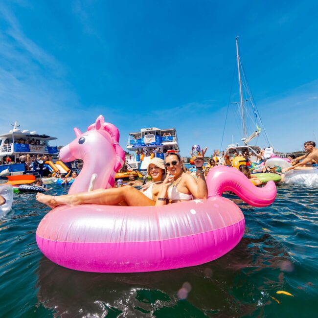 People are enjoying a sunny day on the water with inflatable floats, including a pink unicorn. Boats and yachts surround them. The sky is clear and blue, creating a festive and lively atmosphere.