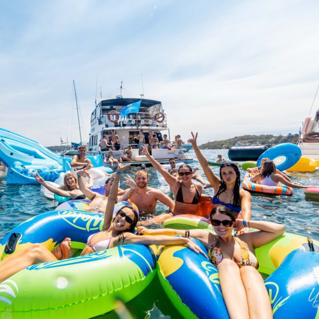 A group of people enjoy a sunny day on the water, lounging on colorful inflatable tubes. Several boats are in the background, and some people are standing or sitting on them. Everyone appears relaxed and cheerful.
