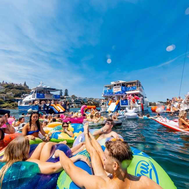 A lively scene on the water features people enjoying a sunny day on colorful inflatable floats. Boats are anchored nearby, and several people are on the decks, socializing. The blue sky and lush greenery frame the vibrant gathering.