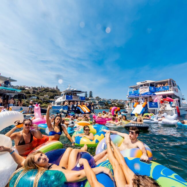 A group of people enjoying a sunny day on colorful inflatable pool floats in the water, with two yachts in the background. The scene is festive, with clear blue skies and everyone appearing relaxed and joyful.