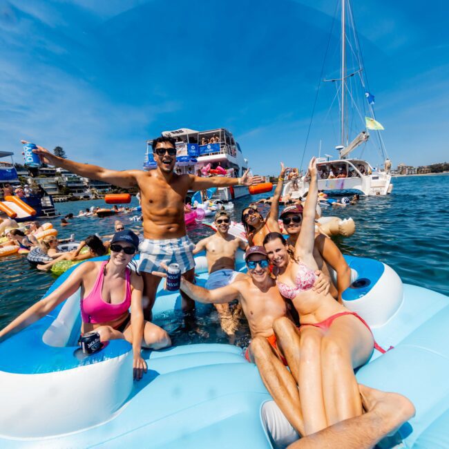 A group of people in swimwear relax and pose on a large inflatable float in the water. Boats and more people on floats are in the background under a bright blue sky. Everyone looks cheerful and festive.
