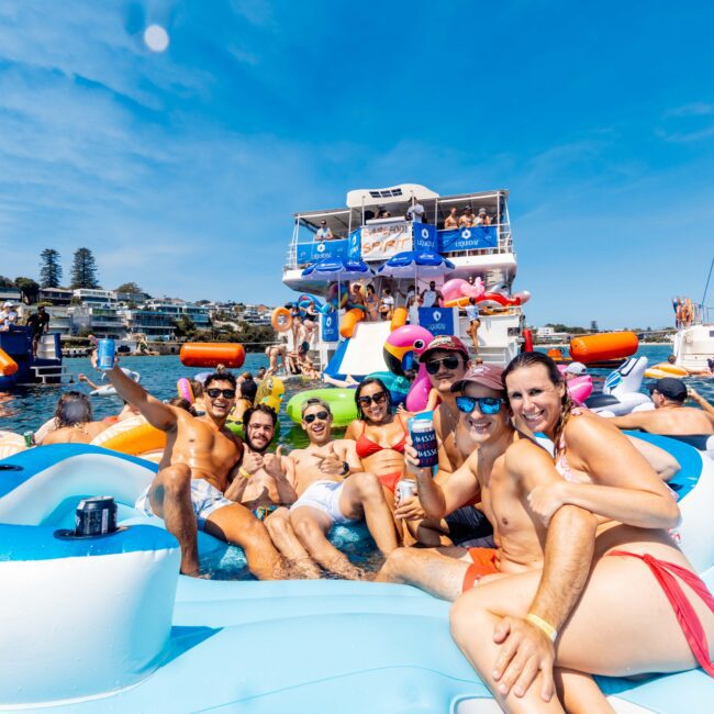 A group of people are enjoying a sunny day on the water, lounging on inflatable floaties. They are smiling and wearing swimsuits, with boats visible in the background. The sky is clear and blue, adding to the lively, festive atmosphere.