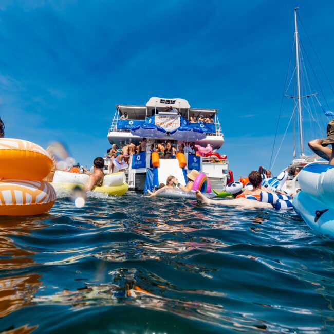 A lively scene of people swimming and floating on inflatables near a large boat in the water. The sky is clear and blue, with a sailboat mast visible on the right. The atmosphere is festive and summery.