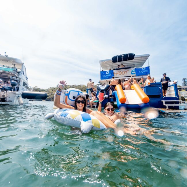 A group of people enjoy a sunny day on the water. Two smiling individuals pose with sunglasses on an inflatable device. In the background are boats and a water slide with people. The scene is festive and joyful.