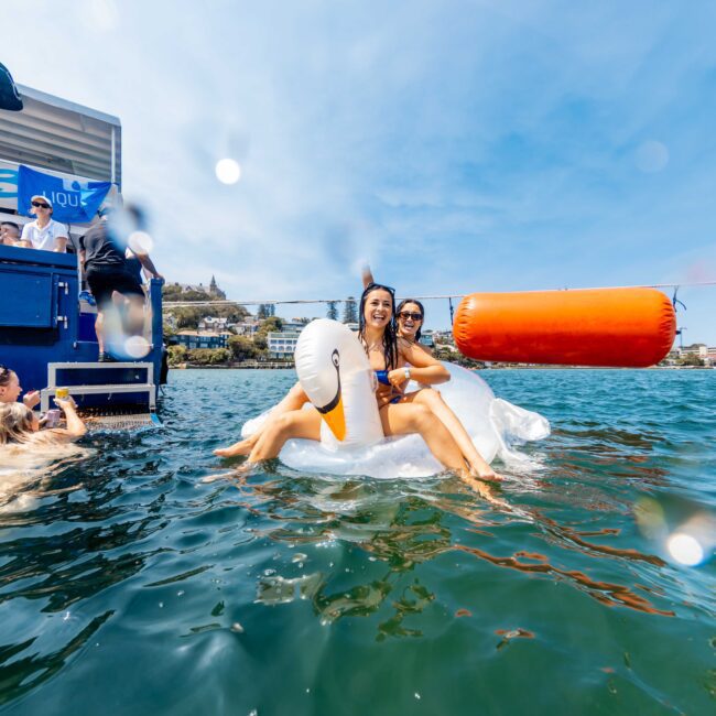 Two women in swimsuits are sitting on a large inflatable swan in the water. They are smiling and surrounded by other people enjoying the sunny day. A boat is on the left, and there’s a buoy in the background.