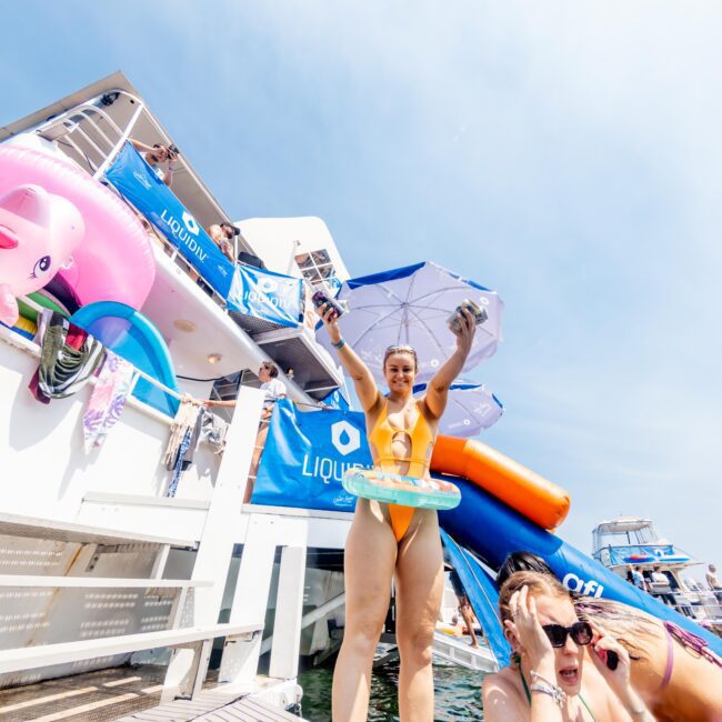 A woman in an orange swimsuit stands on a dock, holding a phone and inflatable toy, with a boat and inflatable flamingo in the background. Two other people are nearby, one with a hand on their head. The scene is sunny and festive.